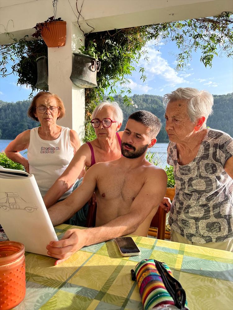 Three women leaning in to look at a sketchbook on a table above the water