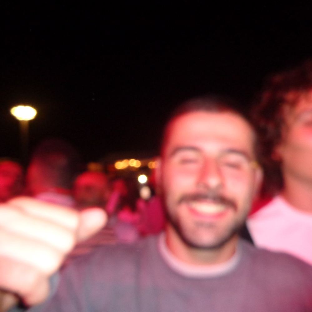 A blurred thumbs-up at night, a crowd and streetlights behind