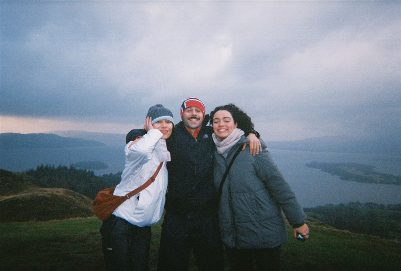 Three friends on a Scottish hilltop above a loch, wrapped up against the cold