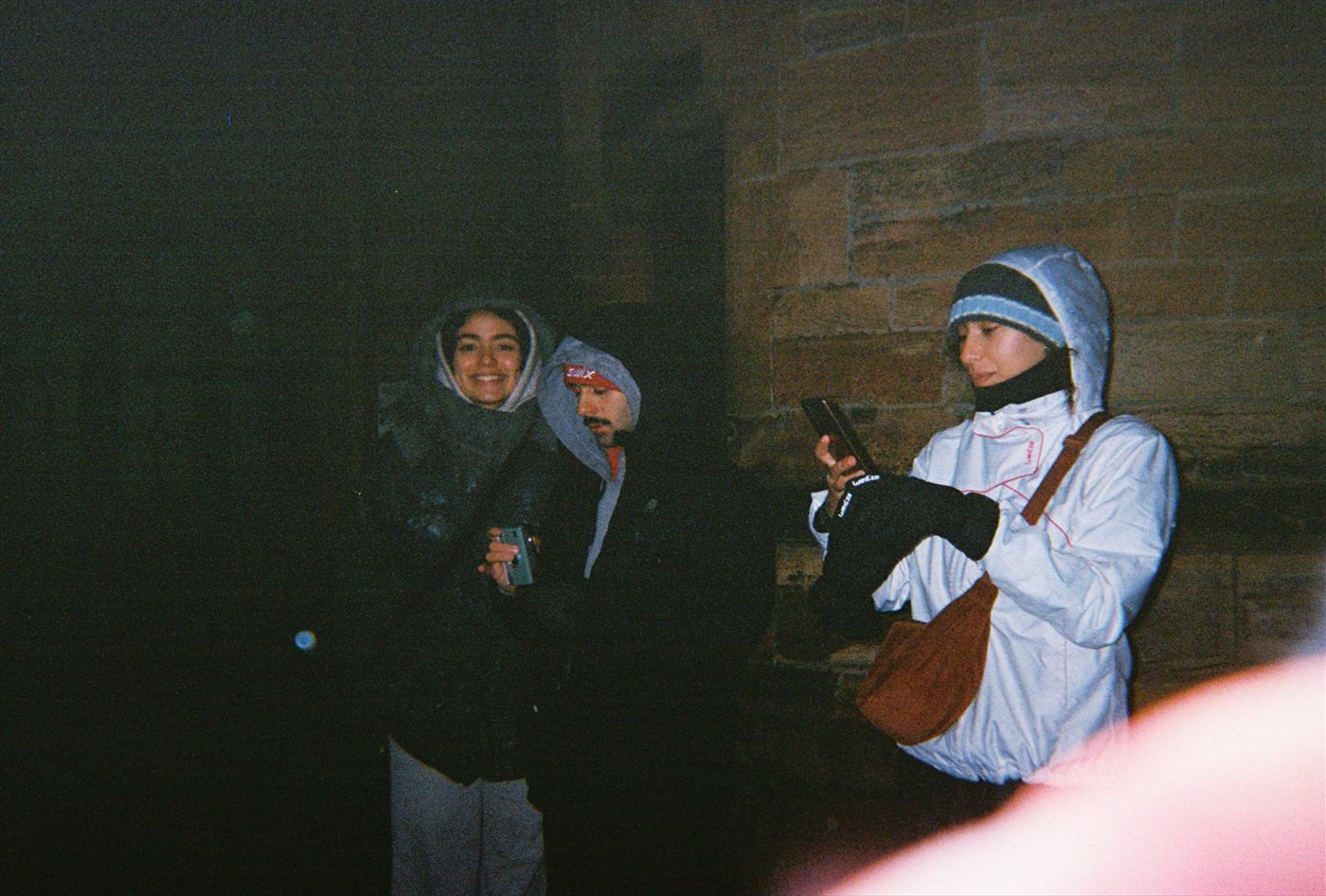 Three friends in winter gear at night against a stone wall