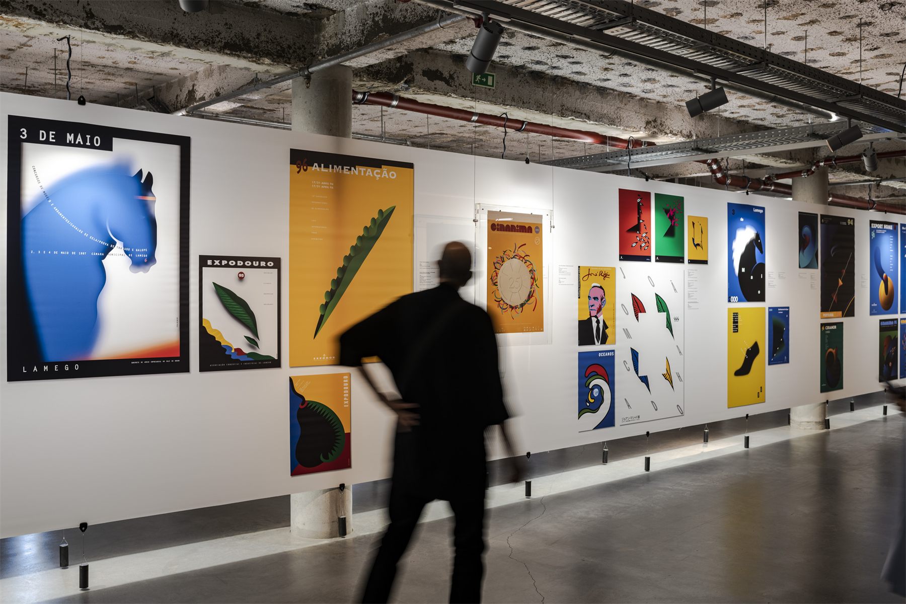 A long white wall of Jo&atilde;o Machado posters running down the gallery, a visitor walking past in motion blur