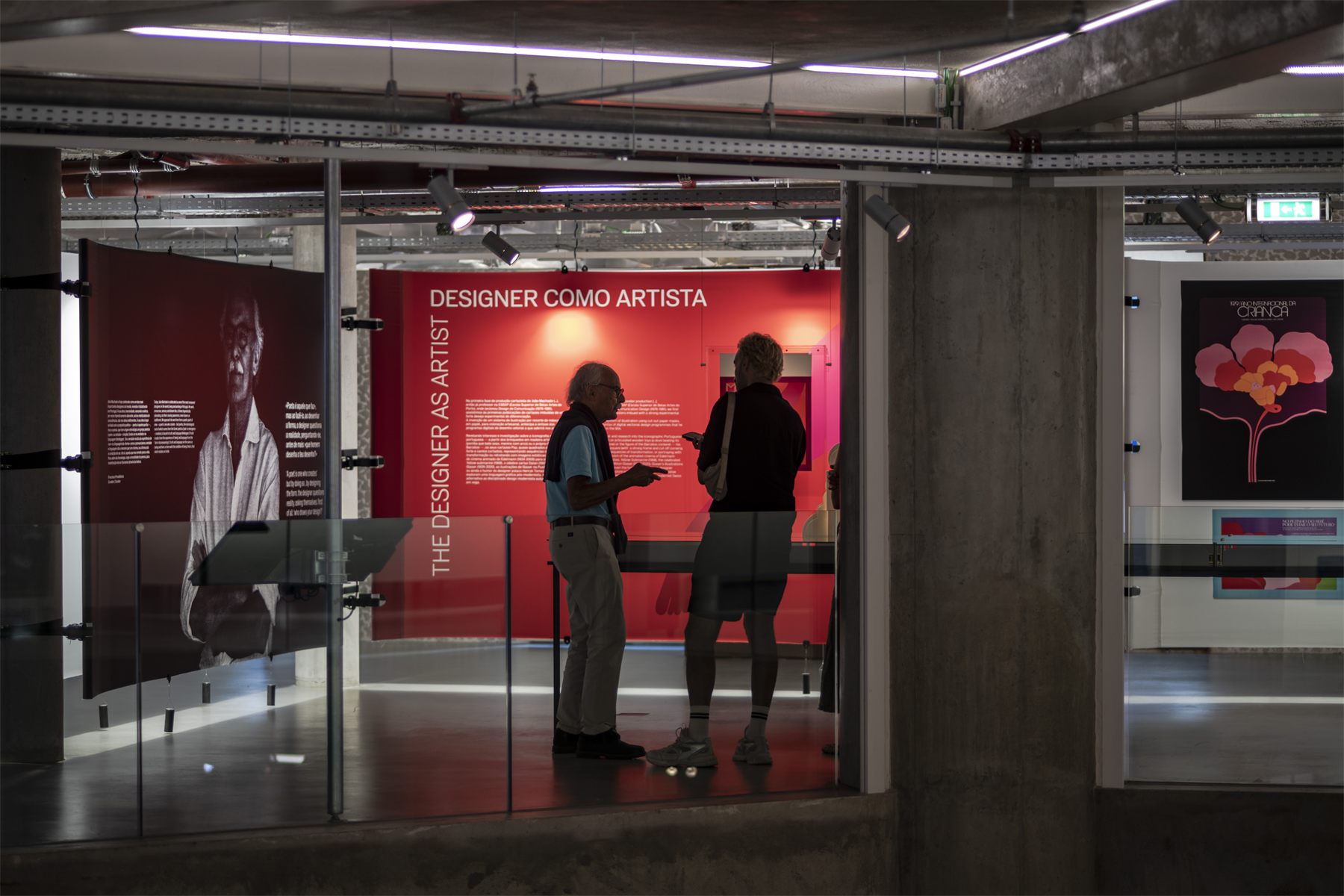 Two visitors talking in front of the red Designer como Artista panel, seen from the corridor through the glass