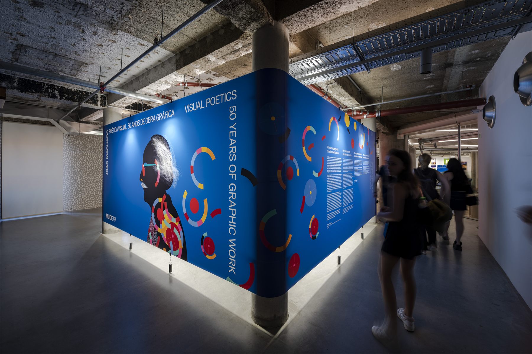 The corner panel built: one continuous blue surface turning around a concrete column, carrying the exhibition title across both faces