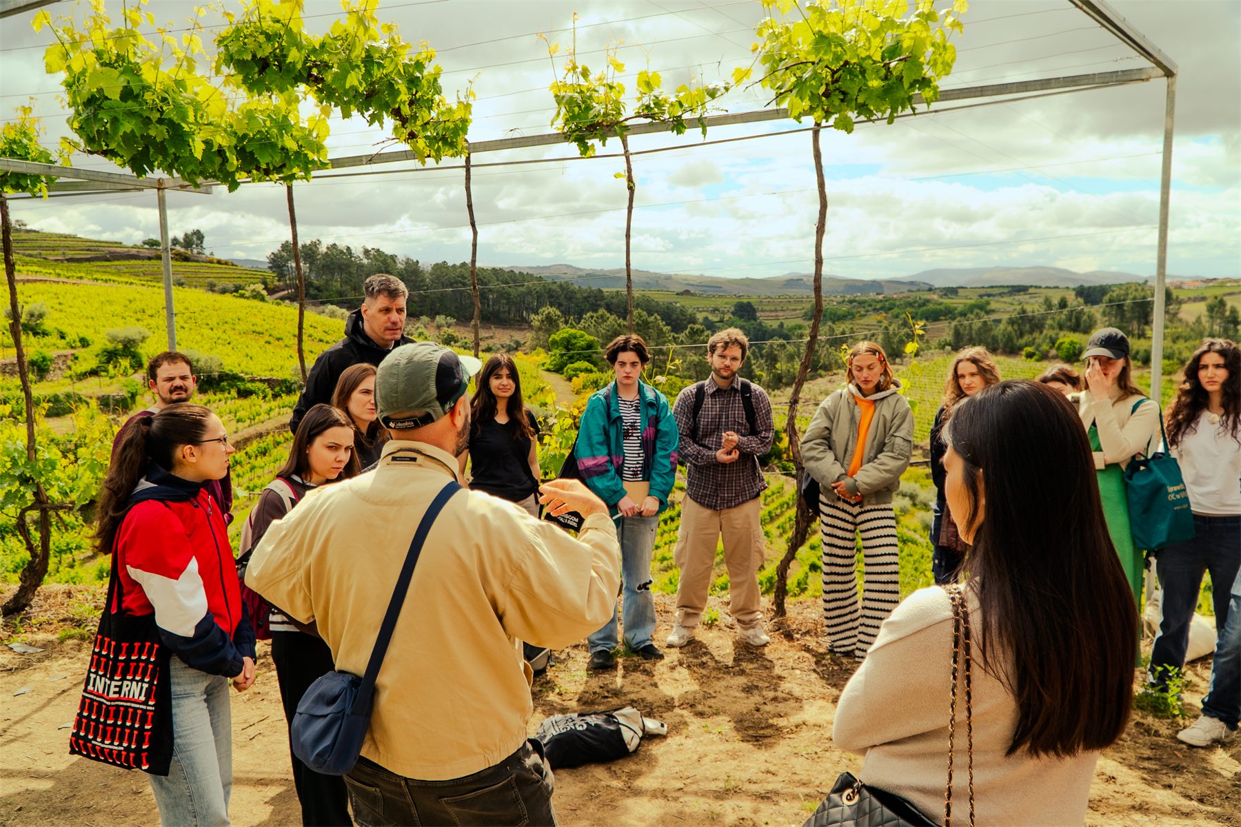 The group standing under a vine pergola, listening, the terraced valley behind them