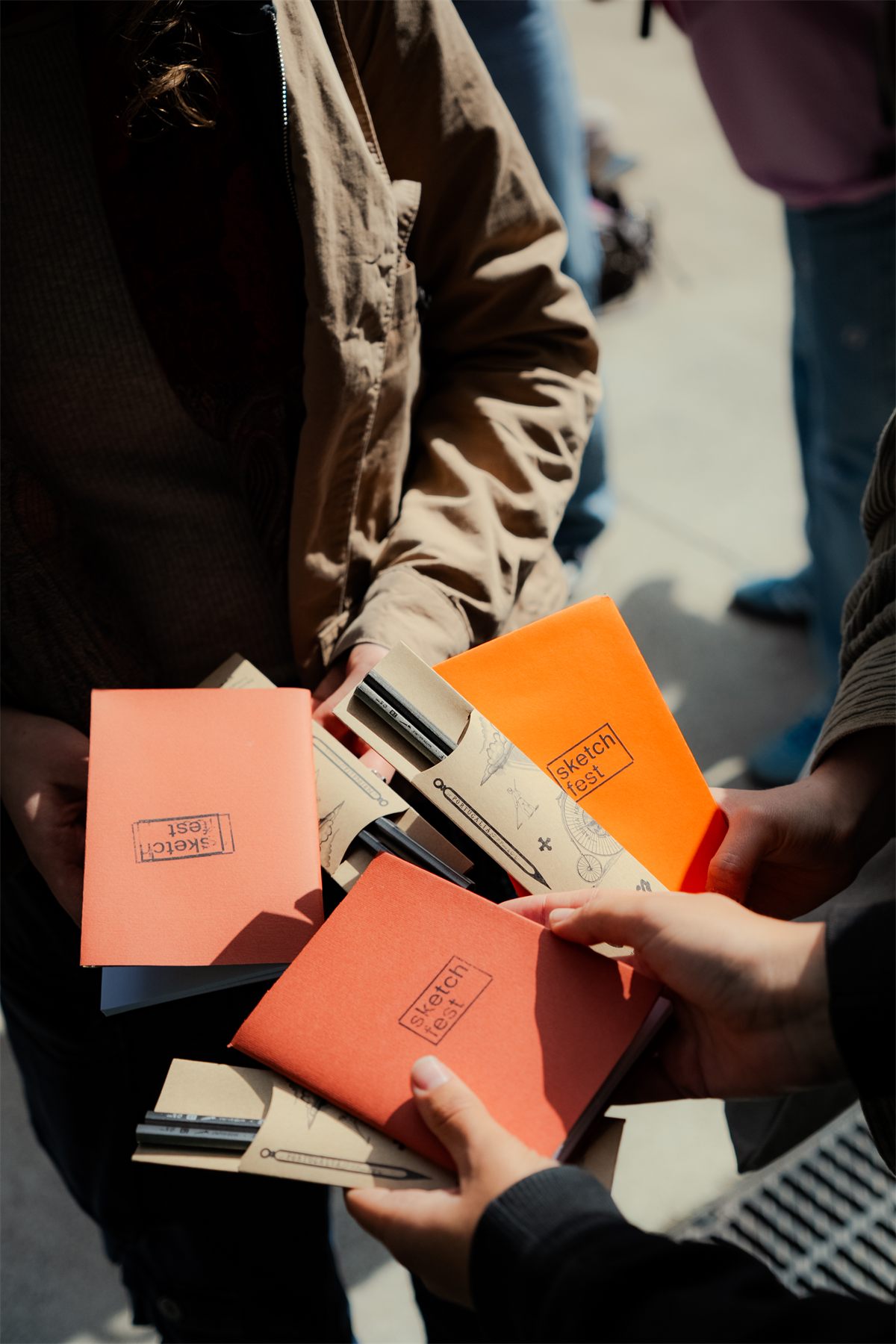 Hands holding several orange Sketch Fest sketchbooks with pencil kits tucked into them