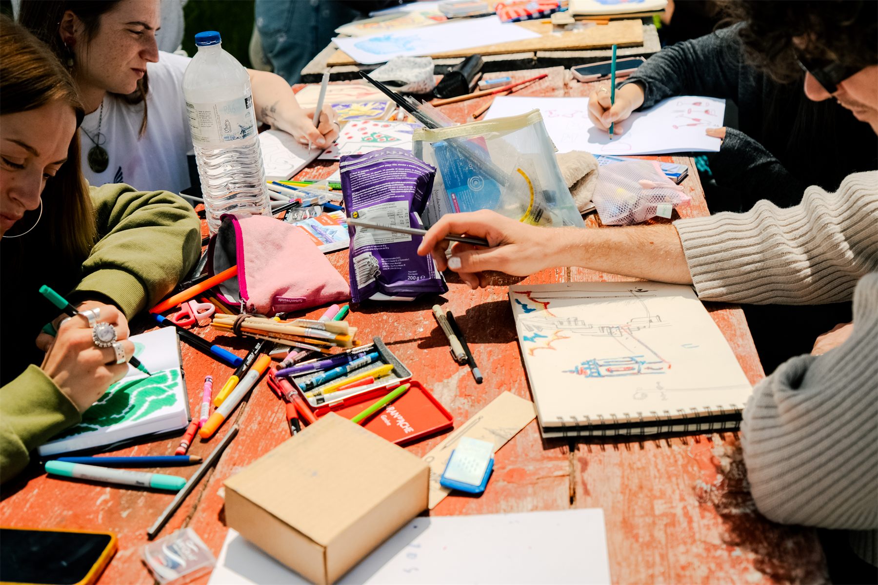 A long table covered in sketchbooks, markers and pencils, several people drawing at once