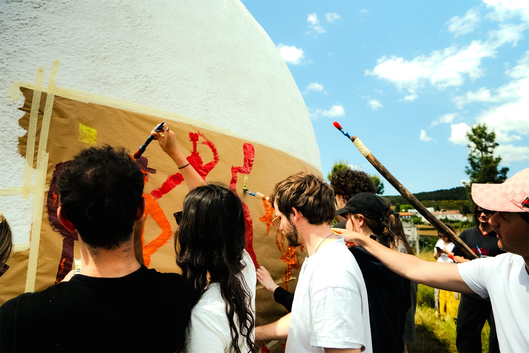 Participants painting a full-size tank wrapped in kraft paper, using long-handled brushes under a blue sky
